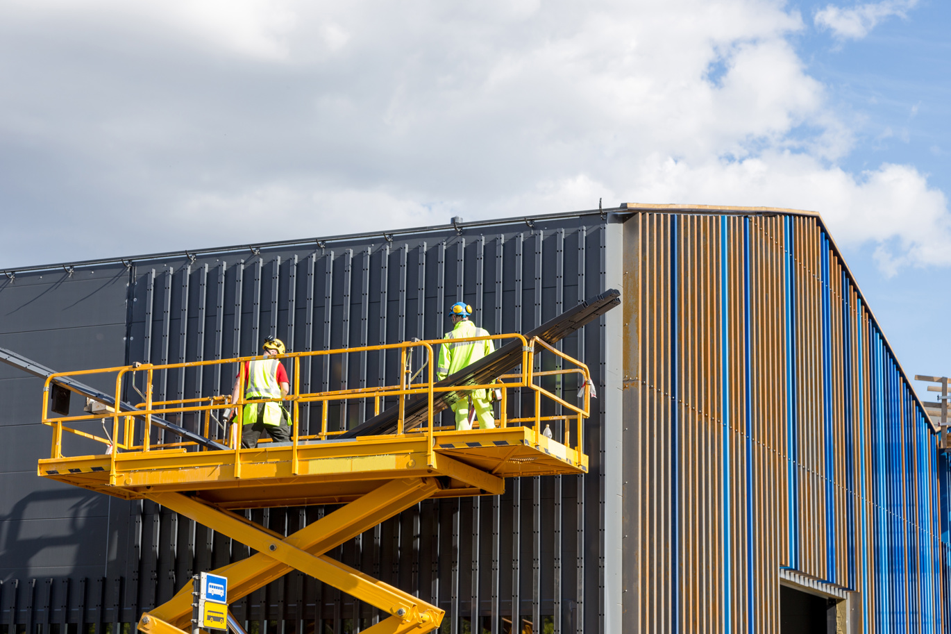 Yellow hydraulic lift with two engineers in high visibility PPE high up next to a warehouse.