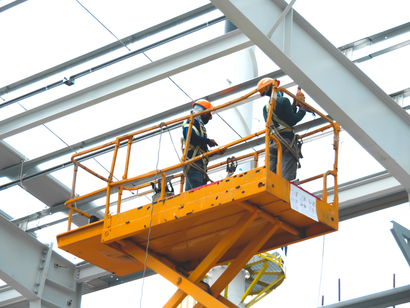 Indoors orange hydraulic lift with two workers high up wearing protective harnesses and orange hard hats.