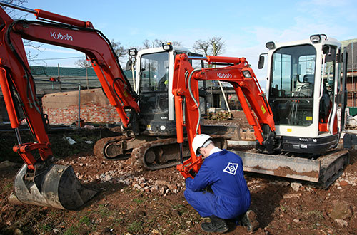 Inspector in a blue jumpsuit and white hard hat crouched down next to two mini excavators on a construction site.